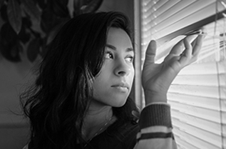 Una chica con pelo oscuro tocando las persianas y mirando por la ventana